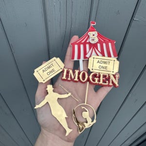 May include: A selection of circus-themed cake toppers. Features a red and white striped circus tent, gold-coloured ticket stubs that read "ADMIT ONE", and gold-coloured silhouettes of a ringmaster and a gymnast. The name "IMOGEN" is also displayed.