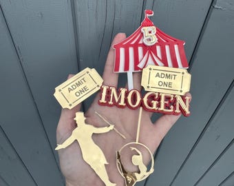 May include: A collection of circus-themed cake toppers. Includes a red and white striped circus tent, gold-colored ticket stubs that read "ADMIT ONE", and a gold-colored silhouette of a ringmaster and a gymnast. The name "IMOGEN" is also featured.