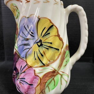 May include: White ceramic pitcher with a floral design featuring purple, yellow, and pink pansies. The pitcher has a ruffled rim and a curved handle. Brown and green leaves accent the flowers.