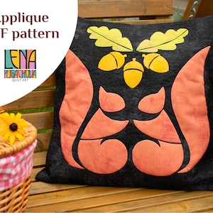 May include: A black pillow with an applique design of two squirrels and acorns. The squirrels are orange and the acorns are yellow. The pillow is on a wooden bench.