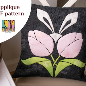Puede incluir: Una almohada negra con una aplicación rosa y verde de dos tulipanes y orejas de conejo. La almohada está sobre una silla blanca con una alfombra verde y blanca en el fondo. El texto "Applique PDF pattern" y "Lena Pugachova Quilt Art" está en la imagen.
