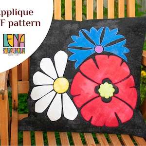 May include: A black pillow with three felt appliques of flowers. The flowers are a white daisy, a red poppy, and a blue cornflower. The pillow is on a wooden chair.