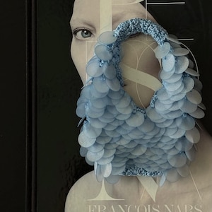 May include: A light blue handbag featuring a crocheted base covered in overlapping circular translucent sequins. The bag has a rounded handle and a textured, layered appearance created by the dense arrangement of the small, round, pale blue plastic discs.