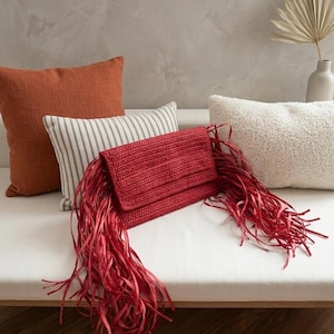 以下が含まれることがあります： 白い表面に、長いフリンジが付いた赤いクラッチバッグが置かれています。オレンジ色とストライプのクッションがそばにあります。クラッチバッグは長方形で、フラップ開閉式です。背景にはニュートラルな壁と装飾要素があります。