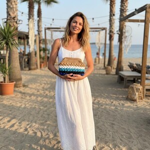 May include: A woman in a white dress holds a handmade clutch. The clutch features a textured design with brown, blue, and white sections. The background includes palm trees, a beach, and a clear sky.