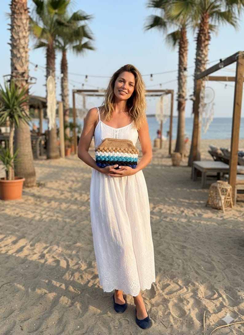 May include: A woman in a white dress holds a handmade clutch. The clutch features a textured design with brown, blue, and white sections. The background includes palm trees, a beach, and a clear sky.