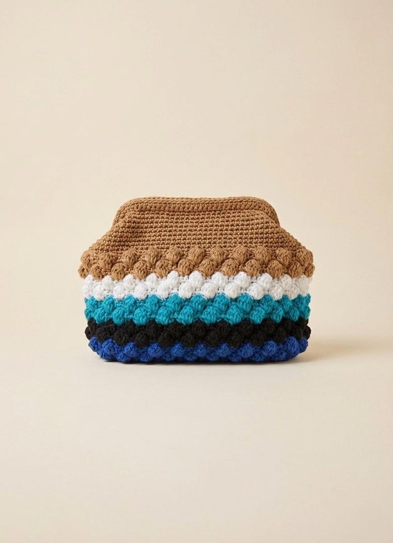 May include: A handmade crochet clutch in a variety of colors. The top is a solid brown, followed by white, blue, black, and blue rows. The clutch has a textured, bobble stitch design.