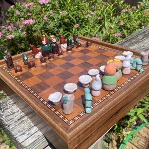 Puede incluir: Un tablero de ajedrez de madera con un patrón de tablero marrón y beige. Las piezas de ajedrez están hechas de cerámica y presentan un tema de jardín caprichoso, que incluye hongos, gnomos y pájaros.