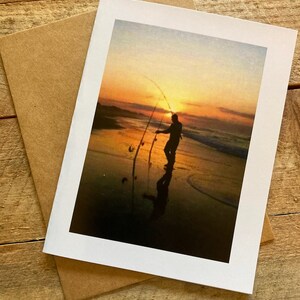 May include: A silhouette of a person fishing at sunset on a beach. The sky is a vibrant orange and yellow, and the water is calm and reflective.