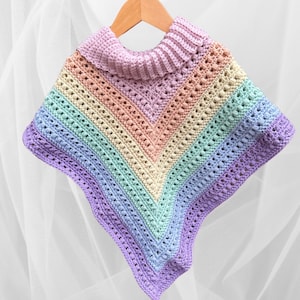 May include: A handmade, rainbow-colored crochet poncho with a ribbed, light purple turtleneck. The poncho features a V-neck design and is suspended on a wooden hanger. The color scheme includes pastel shades of pink, peach, yellow, green, and purple.