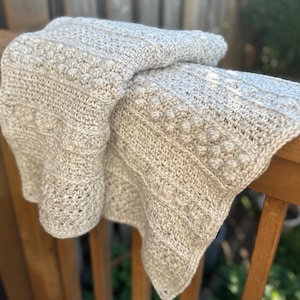 Puede incluir: Una manta de ganchillo gris claro con un patrón texturizado. La manta presenta filas de bolitas y rayas verticales, sobre una barandilla de madera. El color suave y neutro y las costuras detalladas crean una estética acogedora y hecha a mano.