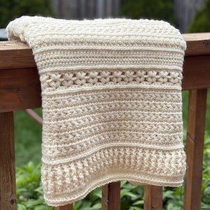 May include: Cream-colored crocheted blanket with intricate stitch patterns. The blanket features textured rows, floral-like designs, and a scalloped edge. The handmade blanket is draped over a wooden railing.
