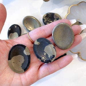 May include: A collection of five oval-shaped pyrite stones, some with a smooth, polished surface and others with a rough, natural texture. The stones are a mix of black and gold colors.