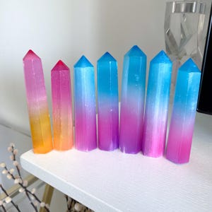 May include: A set of eight ombre crystal points in shades of pink, blue, and yellow. The points are arranged in a row on a white surface.