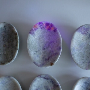 May include: Oval-shaped stones with a marbled appearance. The stones have a base color of white and gray, with patches of vibrant purple and orange hues. The stones are arranged on a light gray surface.