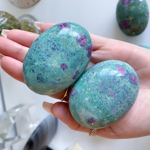 May include: Two oval-shaped gemstones with a teal base color and scattered red inclusions. The stones are smooth and polished, held in a hand. The gemstones have a natural, organic appearance, suggesting they are crystals or minerals.