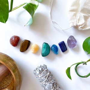 May include: A set of seven chakra crystals, including red jasper, tiger's eye, citrine, green aventurine, blue lace agate, lapis lazuli, and amethyst, arranged in a rainbow pattern on a white surface. A white sage smudge stick and a brass singing bowl are also visible in the image.