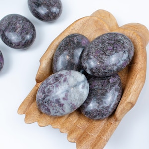 May include: Oval-shaped stones with a mix of dark purple and gray hues, some with flecks of lighter purple. Several stones are nestled in a carved wooden bowl, with others scattered nearby. The stones have a polished, smooth appearance.