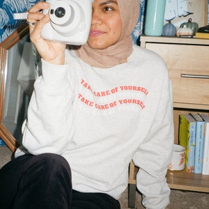 May include: A person wearing a beige headscarf and a grey sweatshirt with the text "Take care of yourself" printed in red. The person is holding a white instant camera in front of their face.