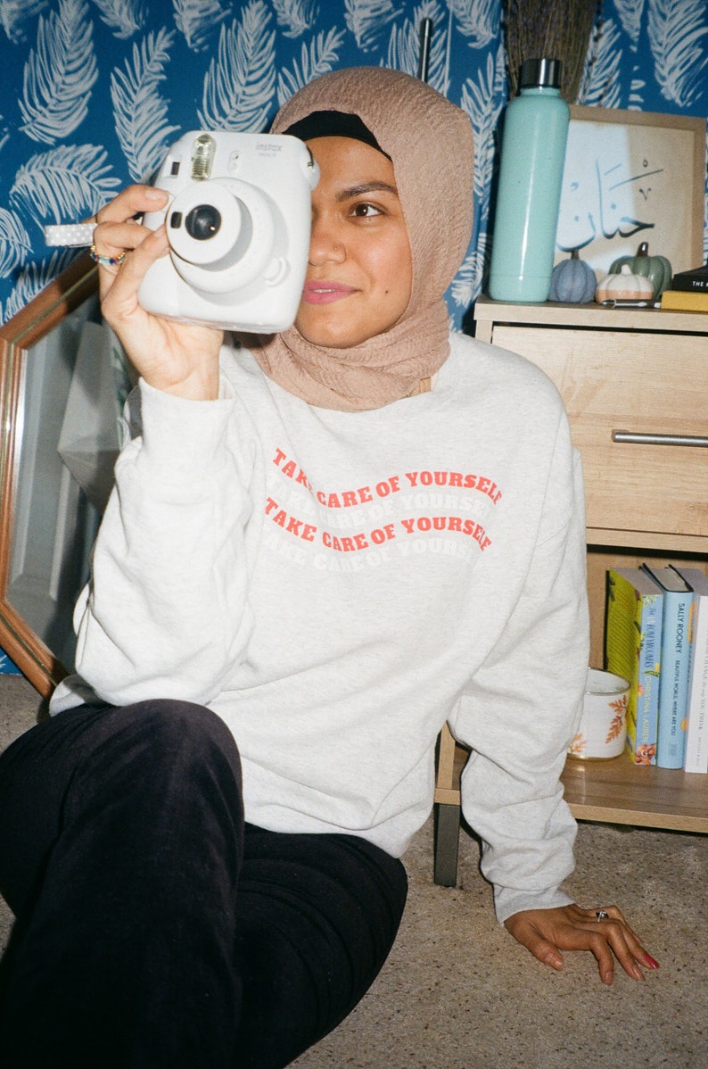 May include: A person wearing a beige headscarf and a grey sweatshirt with the text "Take care of yourself" printed in red. The person is holding a white instant camera in front of their face.