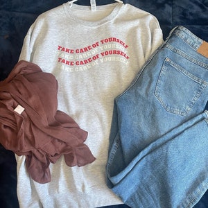 May include: A grey sweatshirt with the phrase "Take care of yourself" repeated in red lettering. The sweatshirt is hanging on a hanger and is paired with blue jeans.