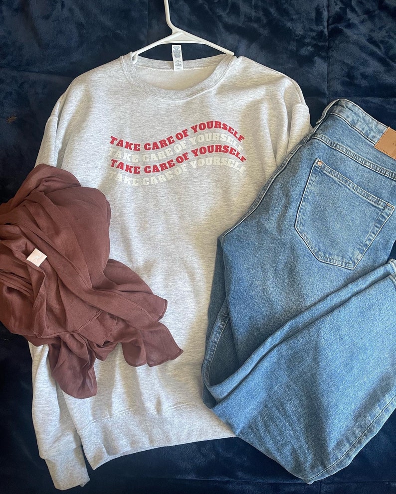 May include: A grey sweatshirt with the phrase "Take care of yourself" repeated in red lettering. The sweatshirt is hanging on a hanger and is paired with blue jeans.