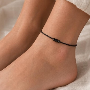 May include: A black anklet with small, dark beads is worn on an ankle. The anklet is made of a thin, black cord. The background is a soft, neutral-toned fabric. A small, beige vase with dried plants is in the background.