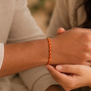 May include: A thin, braided wristband featuring a vibrant, repeating pattern of orange and yellow twisted threads. The cord is tied securely around the wrist, showcasing a simple, minimalist design with bright, warm colors and a smooth, uniform texture.