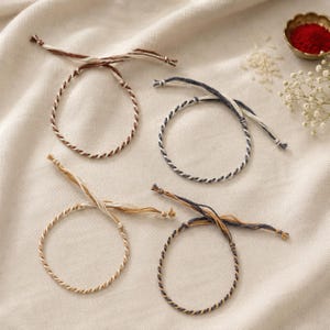 May include: Four thin bracelets made of twisted cord. One is brown and cream, one is navy blue and cream, one is tan and cream, and one is navy blue and tan. Each bracelet features loose, untwisted cord ends for tying, creating a simple circular loop.