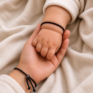 Könnte beinhalten: Nahaufnahme einer Babyhand, die in der Hand eines Erwachsenen ruht. Beide Handgelenke haben schwarze geflochtene Armbänder. Der Hintergrund ist ein weicher, neutralfarbener Stoff. Die Armbänder scheinen verstellbar zu sein.