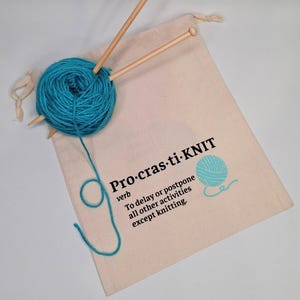 May include: A white canvas bag with a drawstring closure. The bag has a blue yarn ball and knitting needles inside. The bag has the text "Pro-cras-ti-KNIT" printed on it, with the definition: "To delay or postpone all other activities except knitting."