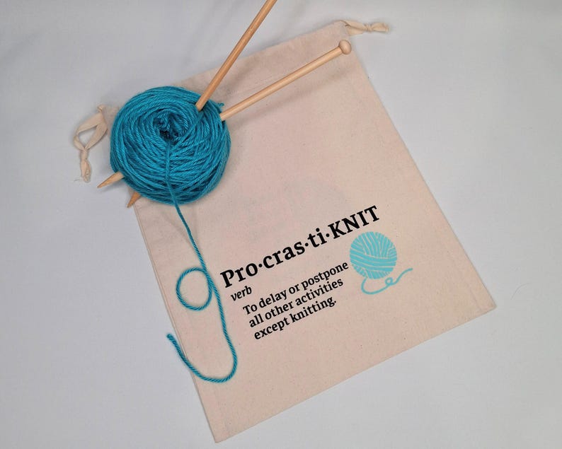 May include: A white canvas bag with a drawstring closure. The bag has a blue yarn ball and knitting needles inside. The bag has the text "Pro-cras-ti-KNIT" printed on it, with the definition: "To delay or postpone all other activities except knitting."