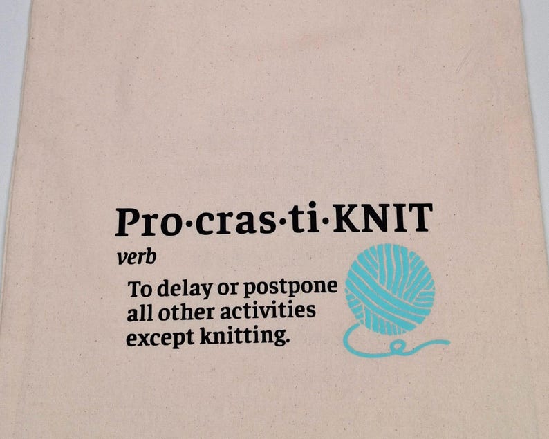 May include: A beige canvas tote bag with black text that reads "Pro-cras-ti-KNIT" and "verb". The text continues with "To delay or postpone all other activities except knitting." A blue yarn ball with a white outline is pictured next to the text.