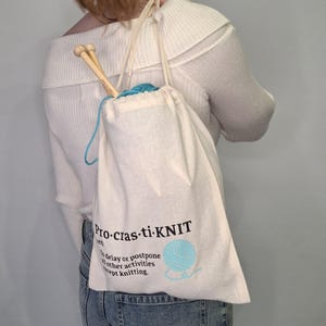 May include: A white canvas drawstring bag with the text "Pro-cras-ti-knit" and the definition "To delay or postpone all other activities except knitting". The bag is filled with blue yarn and knitting needles.