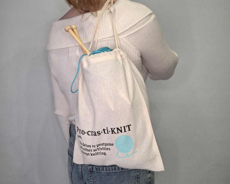 May include: A white canvas drawstring bag with the text "Pro-cras-ti-knit" and the definition "To delay or postpone all other activities except knitting". The bag is filled with blue yarn and knitting needles.