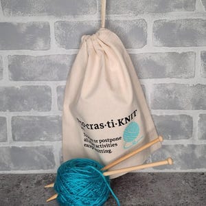 May include: A white drawstring bag with the text "Procrastin-Knit" and a blue yarn ball graphic. The bag is filled with blue yarn and knitting needles.