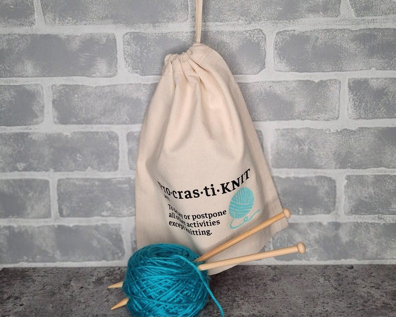 May include: A white drawstring bag with the text "Procrastin-Knit" and a blue yarn ball graphic. The bag is filled with blue yarn and knitting needles.