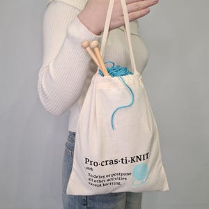 May include: A white canvas tote bag with the text "Pro-cras-ti-KNIT" and the definition "To delay or postpone all other activities except knitting." The bag contains a ball of blue yarn and knitting needles.