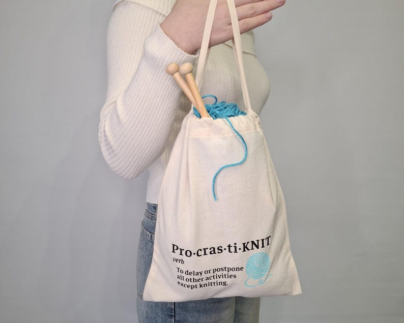 May include: A white canvas tote bag with the text "Pro-cras-ti-KNIT" and the definition "To delay or postpone all other activities except knitting." The bag contains a ball of blue yarn and knitting needles.