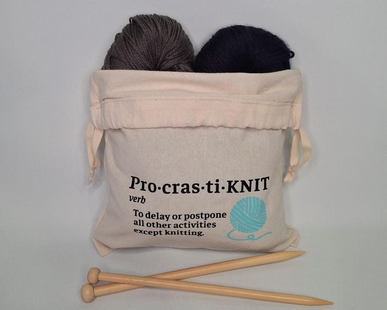 May include: A white canvas drawstring bag with the text "Pro-cras-ti-KNIT" and the definition "To delay or postpone all other activities except knitting."  The bag contains two balls of yarn, one gray and one black, and two wooden knitting needles.