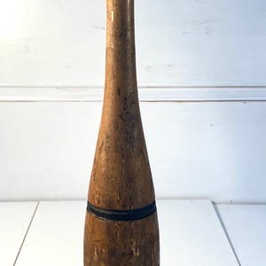 May include: A vintage wooden exercise club with a natural light brown finish, featuring a rounded top knob painted in black and two horizontal black bands encircling the lower body. The wood surface shows signs of wear and texture throughout.