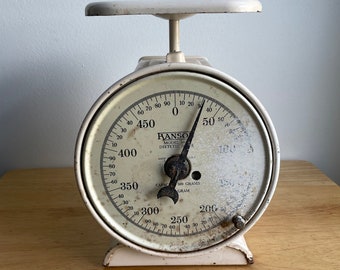 May include: White vintage kitchen scale with a round dial and a black needle. The dial has markings for grams and ounces. The scale is labeled "Ranso Model 115 Dietetic".