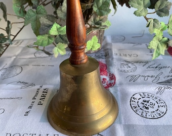 May include: A brass bell with a wooden handle. The bell is sitting on a white fabric surface with black and red stamps.