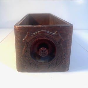 May include: A rectangular dark brown wooden drawer featuring a circular front panel with an embossed leaf-like border and a small, centered round knob. The drawer is open at the top, revealing a hollow interior with visible wood grain texture.