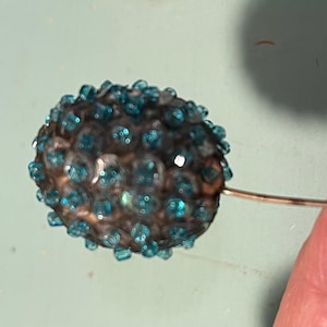 May include: A vintage hat pin with a decorative head. The head is a dark, metallic color and covered in small, teal-colored rhinestones. The pin is held by a hand against a light blue background.