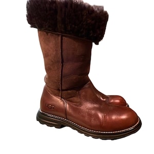 May include: A mid-calf brown leather boot featuring a smooth, polished leather lower section and a soft, dark brown shearling cuff at the top. The boot includes a thick, dark brown rubber sole with visible stitching and the UGG logo embossed on the side.