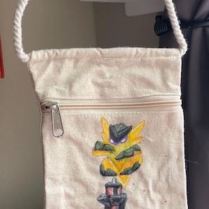 May include: A beige canvas crossbody bag with a zipper closure and a white rope strap. The bag features a hand-painted design of a yellow and black creature on a black and red structure, with red flowers at the base.