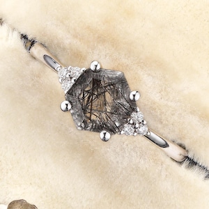 May include: A silver-toned ring featuring a central pentagonal rutilated quartz gemstone with visible dark needle-like inclusions. The stone is flanked by three small clear accent stones on each side, set on a thin, polished metal band.