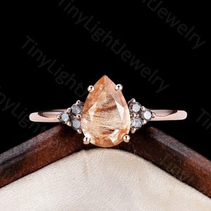 May include: A rose gold ring featuring a pear-shaped peach-colored gemstone at the center, flanked by three small dark gray stones on each side. The ring is set against a dark background with the repeating text TinyLightJewelry visible.