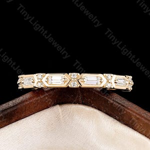 May include: A gold band ring featuring a repeating pattern of baguette-cut diamonds set within hexagonal frames, alternating with small round accent diamonds. The ring is displayed horizontally, showcasing its intricate metalwork and clear stones.